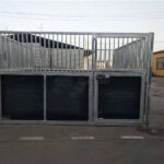 Metal horse stall with a sliding door, featuring vertical bars and dark panels, positioned outdoors.