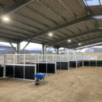 A row of metal-framed stalls with wire mesh and dark panels, set under a large open-sided shed with a corrugated roof. Several people and a wheelbarrow are present in the background, with a rural landscape visible.