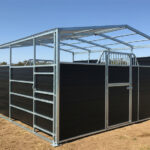 A black horse stall with a metal frame and roof, featuring a partially open front, set in an outdoor area with a clear blue sky in the background.