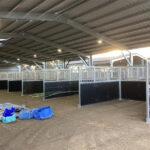 A row of metal-framed horse stalls with wire mesh upper sections and dark lower panels, situated inside a large industrial building with a high ceiling and scattered construction materials in the background.