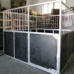 A metal horse stall under construction, with dark panels and vertical bars, located indoors with various supplies in the background.