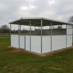 A white horse stall with a roof, featuring vertical bars and dark panels, situated in a grassy field under a cloudy sky.