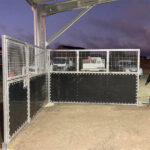 A row of portable horse stables with metal mesh upper sections and black solid lower panels, installed under a metal roof in a barn. The stables are aligned in a straight line, with a concrete floor and a small building with a red roof visible in the background.