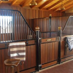 An interior view of a horse barn with several stalls, featuring wooden paneling and black metal bars, showcasing a well-lit and spacious design.
