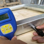 A handheld digital thickness gauge measuring the thickness of a metal frame. The display shows a reading of 84.5 micrometers, indicating the measurement taken.