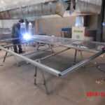 Two workers engaged in welding a metal frame in an industrial workshop, with sparks flying from the welding process and metal components being assembled on a workbench.