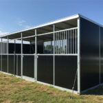 A black metal horse stall with a roof, designed for outdoor use. The structure has multiple stalls and is situated in a grassy area, with a clear blue sky in the background.