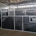 A partially assembled horse stall featuring metal framing and a black panel design, with an open area for feeding visible on one side, set in a warehouse environment.
