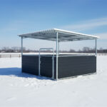 A black horse shelter with a metal roof, featuring an open design and a spacious interior, illuminated by sunlight.
