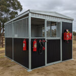 A metal horse stall with a black and silver design, equipped with fire extinguishers and a halter, is situated outdoors with a fenced area in the background.