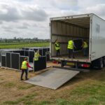 A white truck is unloading black horse stall components, with several workers in fluorescent yellow safety vests busy moving the materials, set against a backdrop of open farmland.