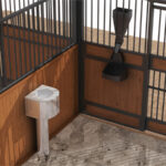 Close-up view of a horse stall corner, showing a water trough and wooden panels with metal grills, designed for comfort and functionality.