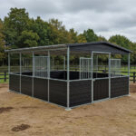 A well-constructed black horse stall with a roof, complete with metal bars for ventilation. The stall is surrounded by grassy terrain and trees, providing a picturesque setting.