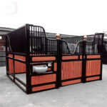 A large horse stall made of black metal and wooden panels, featuring a curved design at the top, with a golden decorative ball on the corners and a white water trough attached to the side.
