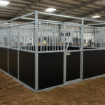 A black horse stall with metal bars at the top, located indoors with a sandy floor and other stalls visible in the background.