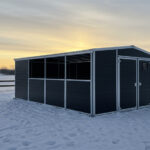 A black horse shelter is set against a winter sunset, with snow covering the ground. The shelter features large windows and a door, and a wooden fence is seen in the background.
