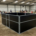 An indoor riding arena with a black horse stall positioned in the foreground. Several riders on horses can be seen practicing in the background.