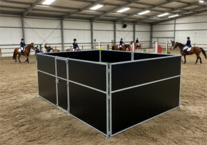 An indoor riding arena with a black horse stall positioned in the foreground. Several riders on horses can be seen practicing in the background.