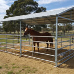 A brown horse standing inside a galvanized steel portable pen with a metal roof for shelter, surrounded by hay on the ground, in an outdoor setting with trees in the background.