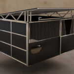 A 3D rendering of a modular horse stable with a metal frame, dark panels, a sloped roof, and a wooden feeder integrated into the design.
