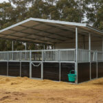 Outdoor horse stable with a galvanized steel frame, metal roof, black panel lower walls, and metal bar upper sections. A green bucket hangs on the wall, and hay is scattered on the ground.