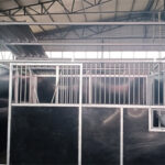 A partially assembled modular horse stable with a metal frame, black panels, and an integrated feeding bowl, situated inside a warehouse.