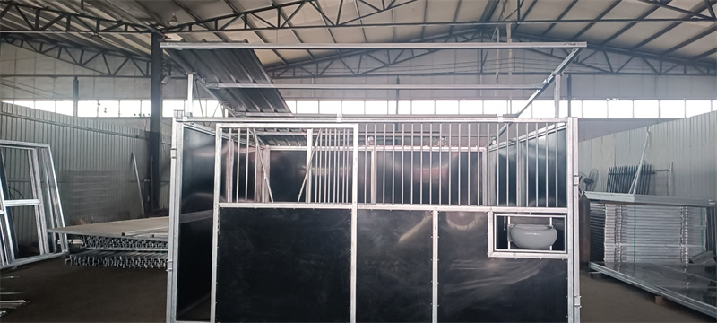 A partially assembled modular horse stable with a metal frame, black panels, and an integrated feeding bowl, situated inside a warehouse.