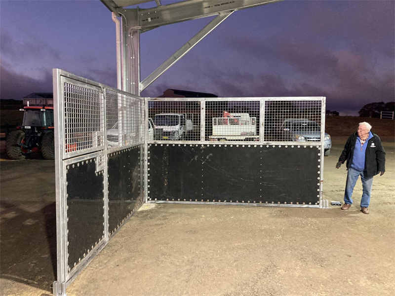 An open-front horse stable or holding pen with metal mesh and black panels, with a man standing nearby in a rural setting at dusk.