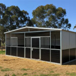 A multi-stall flat-pack horse stable with grey upper metal panels and dark brown lower panels. It features several stalls, one with a dark brown door, and has a metal roof structure.