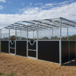 A three-stall DIY horse stable kit with dark brown lower panels, metal bar upper sections, and a translucent corrugated plastic roof. Each stall has a metal gate and a V-shaped opening for the horse's head. The stable is on a dirt and straw base.