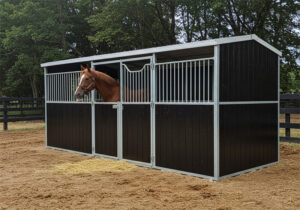 A compact, dark-paneled portable horse stable with silver metal trim and a white roof, featuring two stalls. A bay horse looks out from one stall opening.