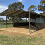 A large open-front DIY horse stable kit with a grey metal roof and grey metal walls, featuring internal metal pipe fencing to create multiple pens. The ground inside is covered with sand or dirt, and the stable sits on a grassy area with trees in the background.