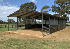 A large open-front DIY horse stable kit with a grey metal roof and grey metal walls, featuring internal metal pipe fencing to create multiple pens. The ground inside is covered with sand or dirt, and the stable sits on a grassy area with trees in the background.