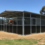 A multi-stall DIY horse stable kit with a grey metal roof and a two-tone design of dark brown lower panels and grey upper sections. A bay horse looks out from one of the internal stalls. The stable is set on a prepared dirt and gravel base.