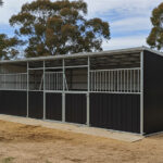 A three-stall DIY horse stable kit with dark brown lower panels and grey metal upper sections and roof. The stalls feature metal bars and gates, and the stable is built on a concrete foundation with straw scattered around it.