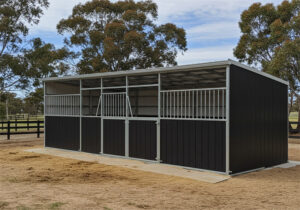 A three-stall DIY horse stable kit with dark brown lower panels and grey metal upper sections and roof. The stalls feature metal bars and gates, and the stable is built on a concrete foundation with straw scattered around it.