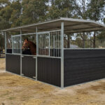 A two-stall flat-pack horse stable with dark brown lower panels, metal bars on the upper sections, and an extended metal roof providing shelter. A bay horse is looking out from one of the stalls.