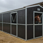 A long, dark-paneled portable horse stable with silver trim and a white roof edge. A bay horse looks out from one of the stable windows.